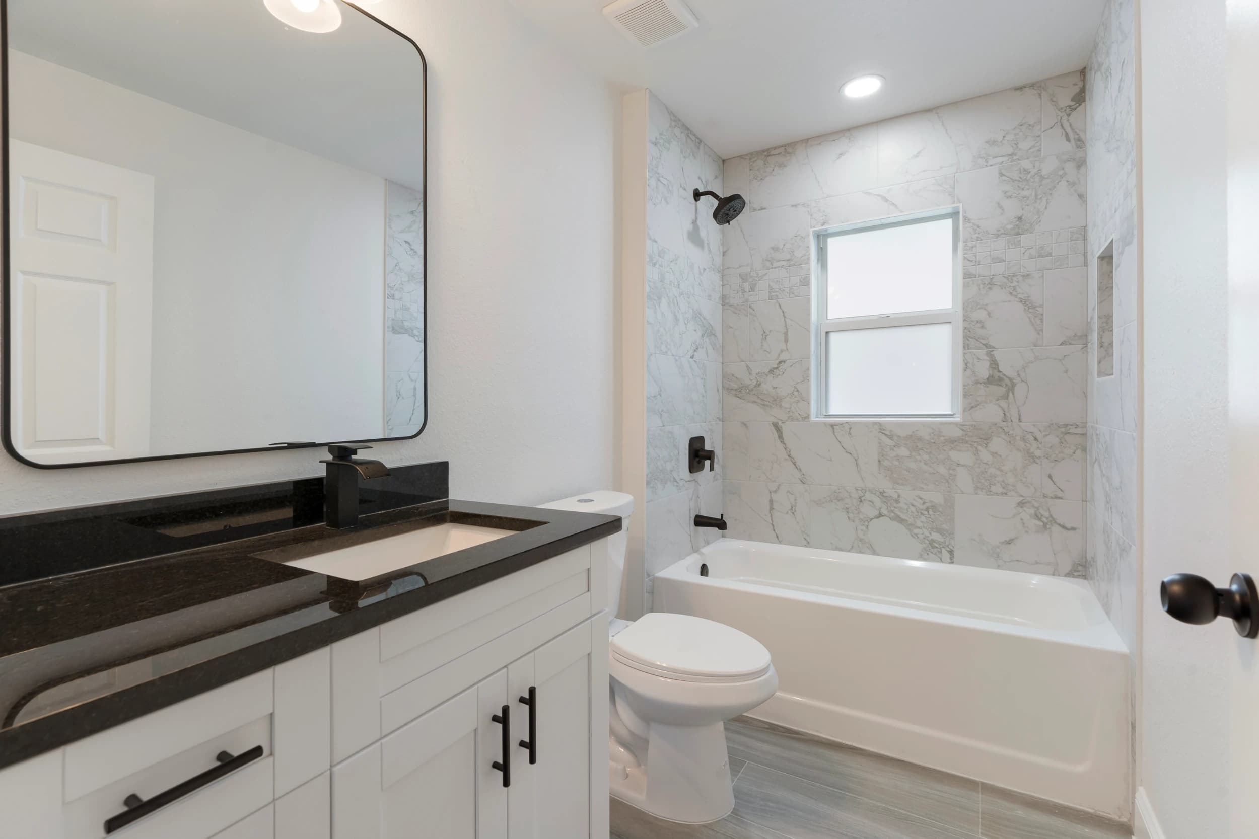 Elegant modern bathroom in a newly constructed home by Vreeland Roofing in Florida. Designed with a sleek black quartz vanity, white shaker cabinets, a spacious bathtub with marble tile surround, and matte black fixtures. A large mirror and natural light from the window enhance the sophisticated and functional design.
