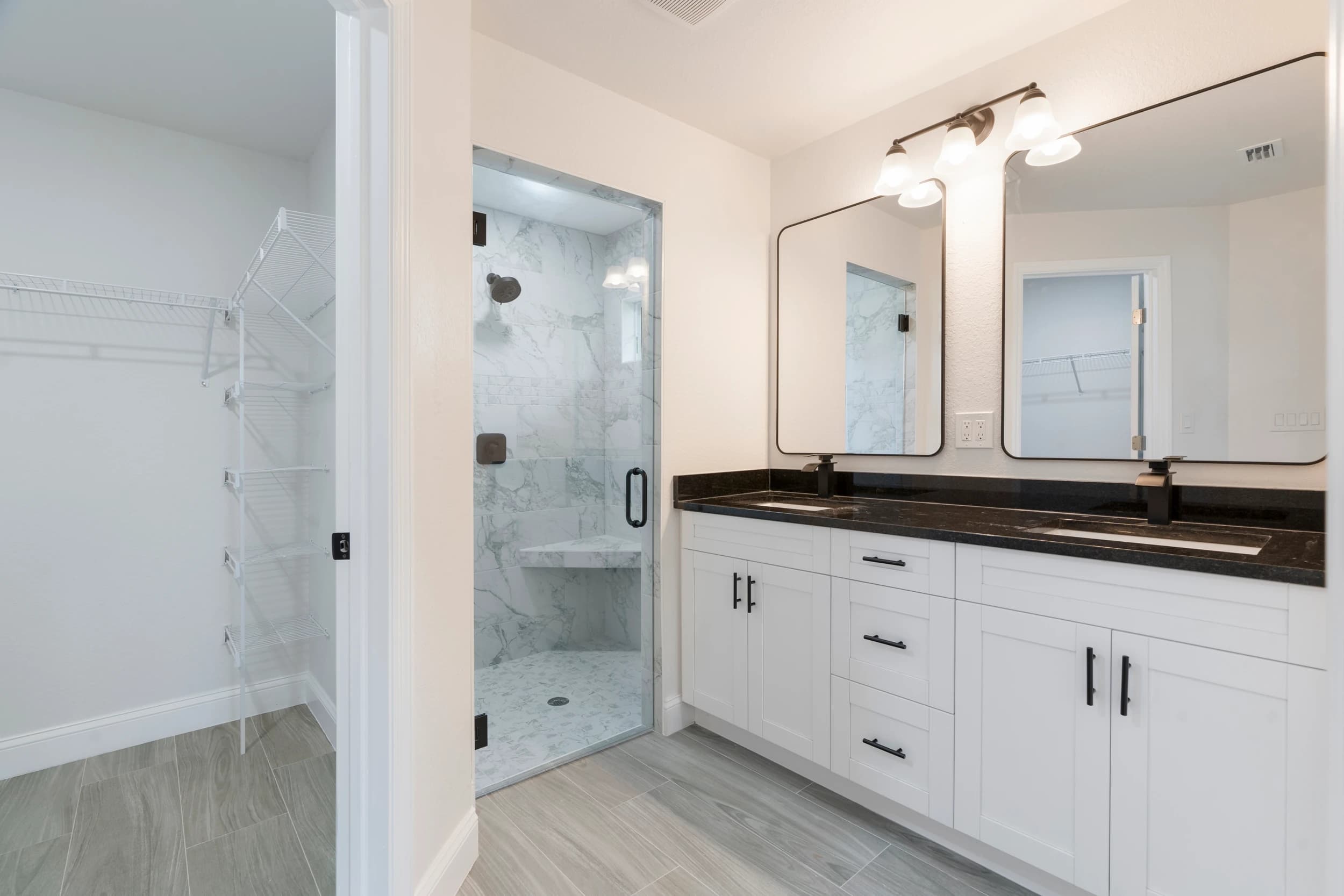 Elegant primary bathroom in a newly constructed Florida home by Vreeland Roofing, featuring a dual vanity with black granite countertops, a frameless glass shower with marble-style tile, and an adjacent spacious walk-in closet with built-in shelving.