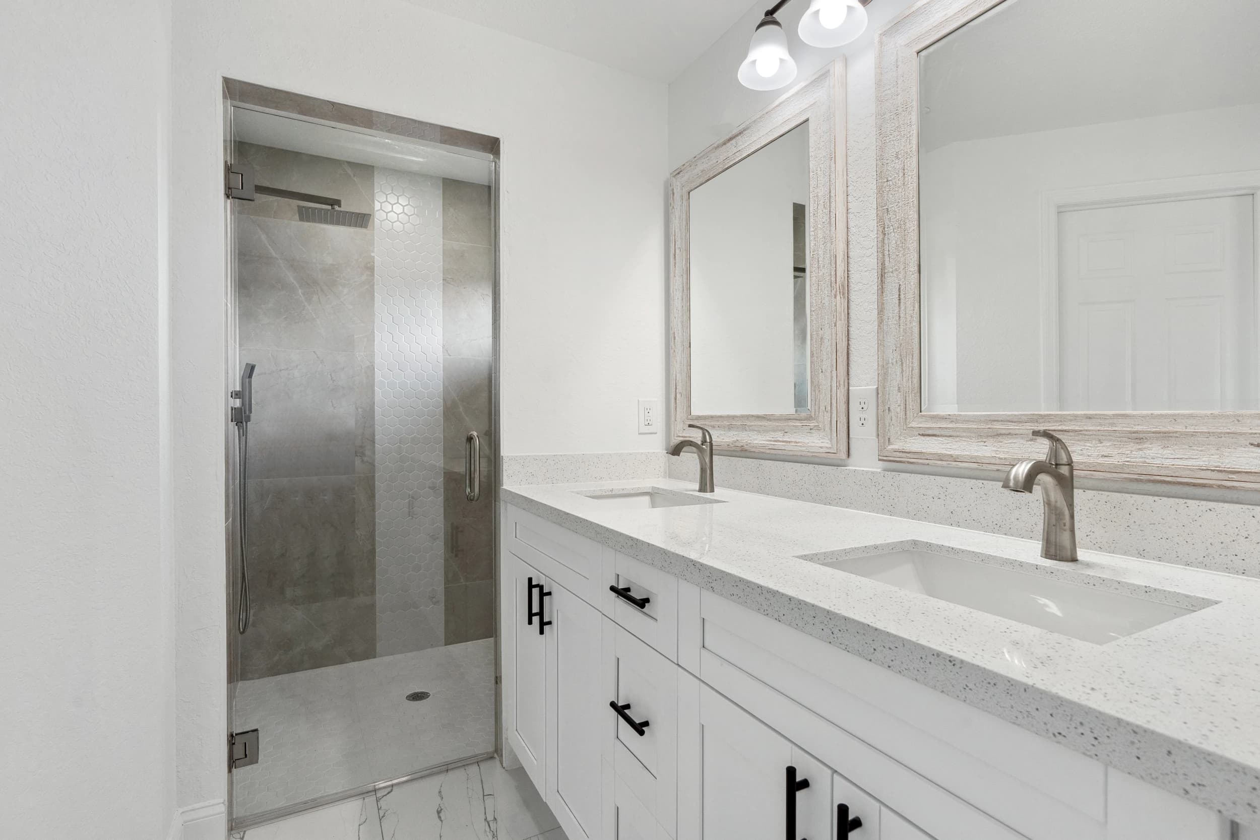 Modern bathroom renovation featuring a double vanity with white quartz countertops, sleek undermount sinks, brushed nickel faucets, and rustic framed mirrors. Walk-in shower with glass doors, hexagonal tile accents, and rainfall showerhead. A Vreeland Roofing home remodeling project in Florida.