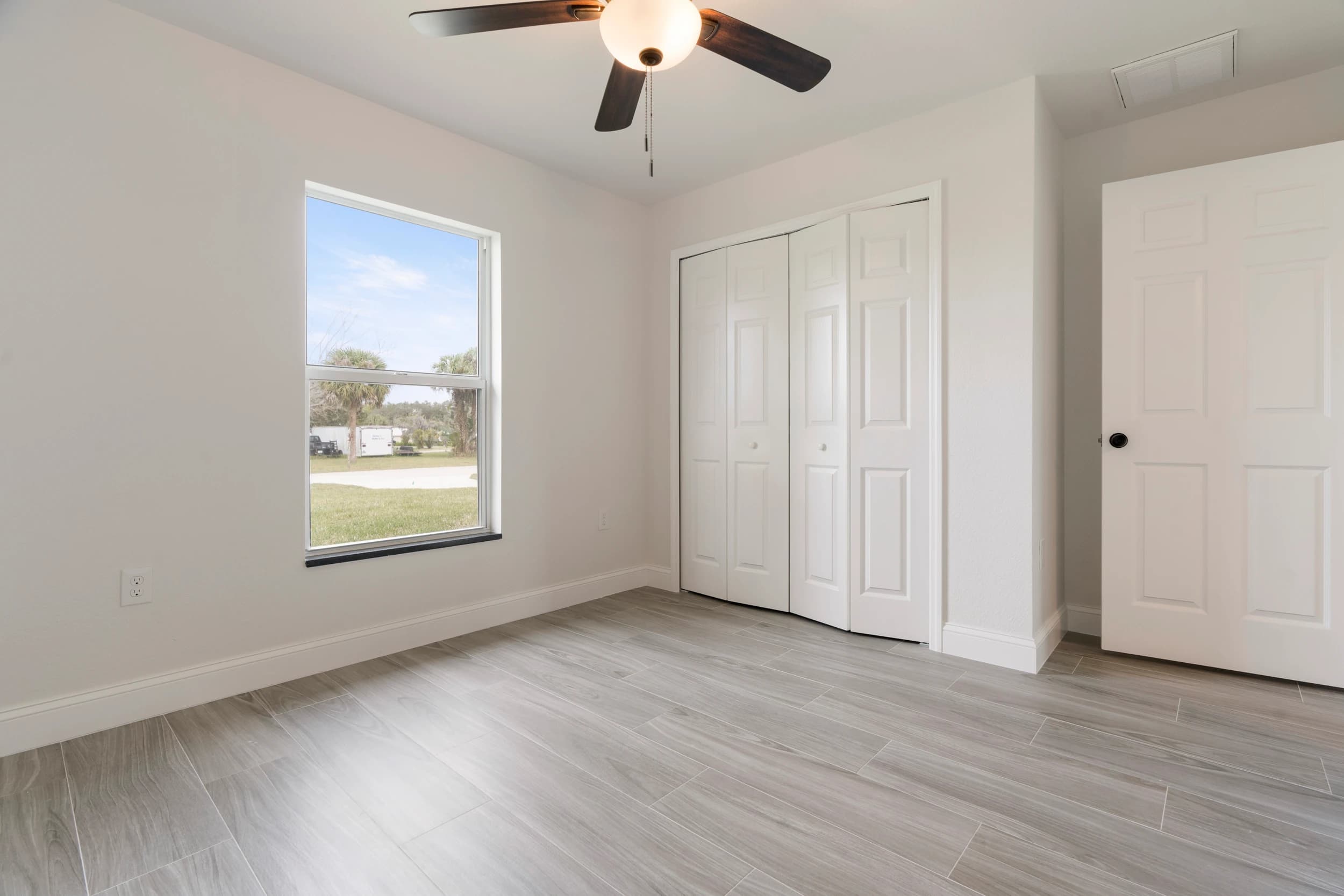 Stylish bedroom with a neutral color palette, large window for natural light, ceiling fan, and built-in closet doors, providing a cozy and modern living space in Florida.