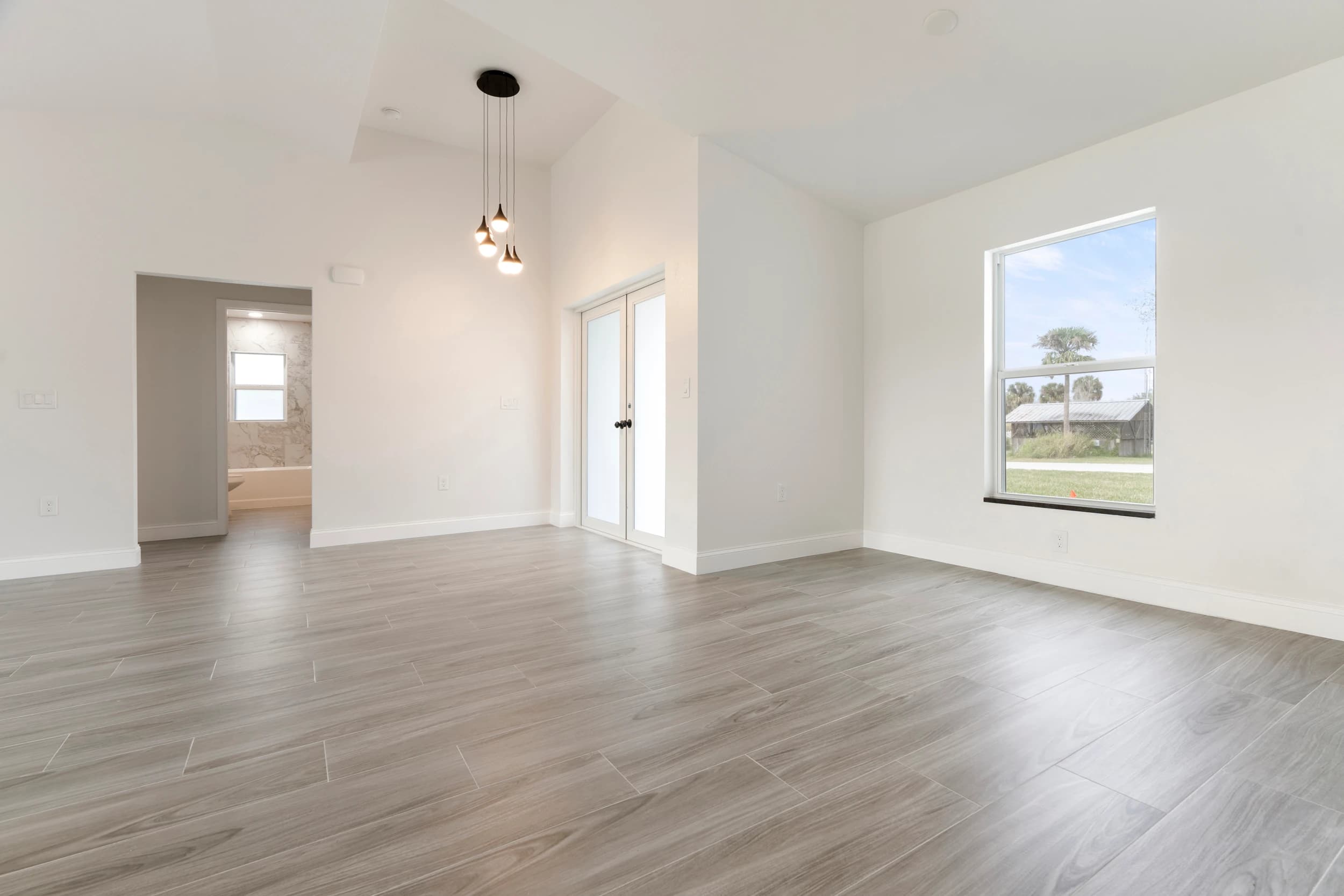 Bright and spacious modern living room in a newly constructed home by Vreeland Roofing in Florida. Featuring high ceilings, elegant wood-look tile flooring, large energy-efficient windows, and a contemporary pendant light fixture. The open-concept design seamlessly connects to other living spaces, enhancing comfort and natural light flow.