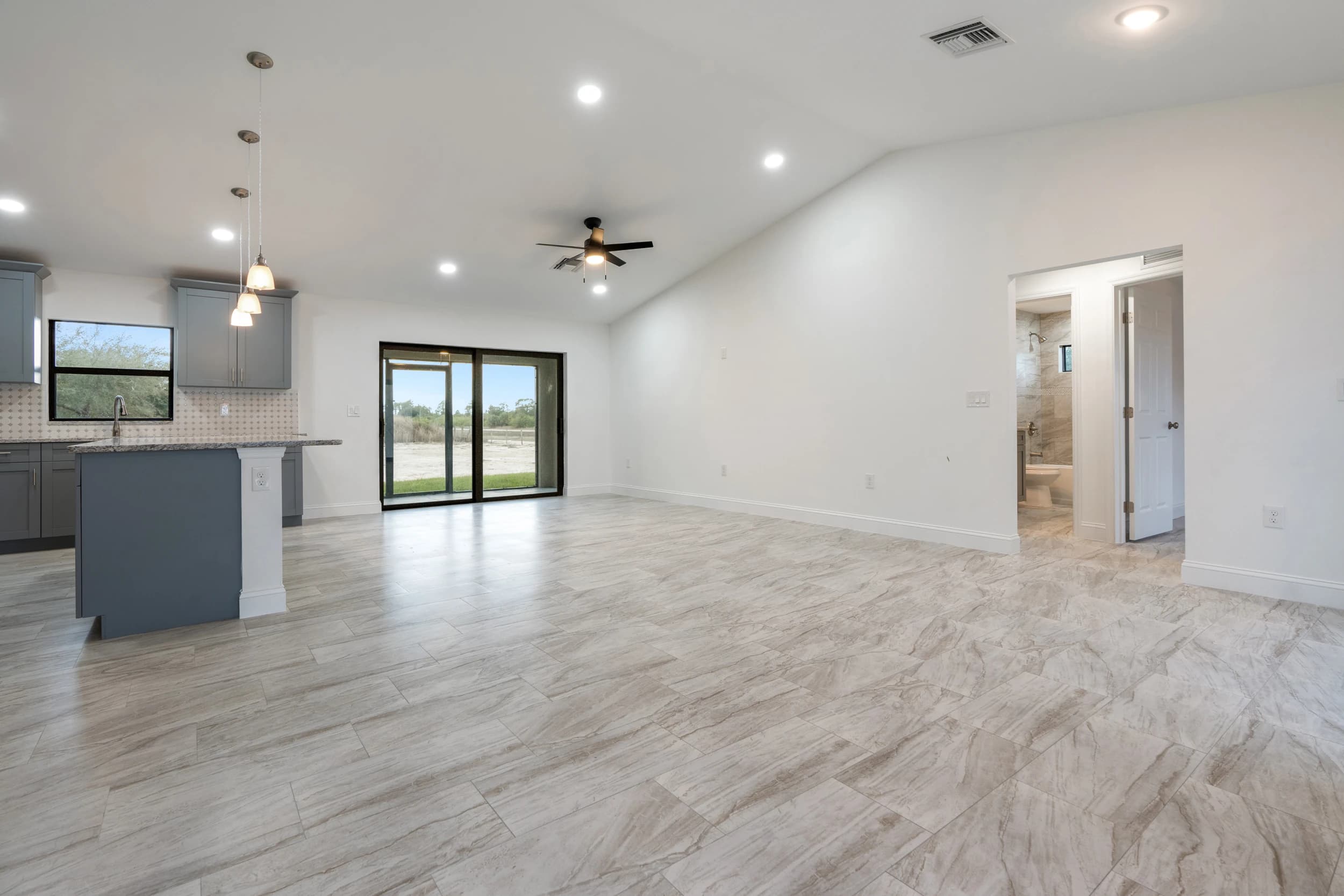 Spacious living room renovation featuring an open-concept layout, high vaulted ceilings, modern recessed lighting, and wood-look porcelain tile flooring. Seamless transition to the kitchen with custom cabinetry and pendant lighting. Large sliding glass doors provide natural light and outdoor access. A Vreeland Roofing home remodeling project in Florida.