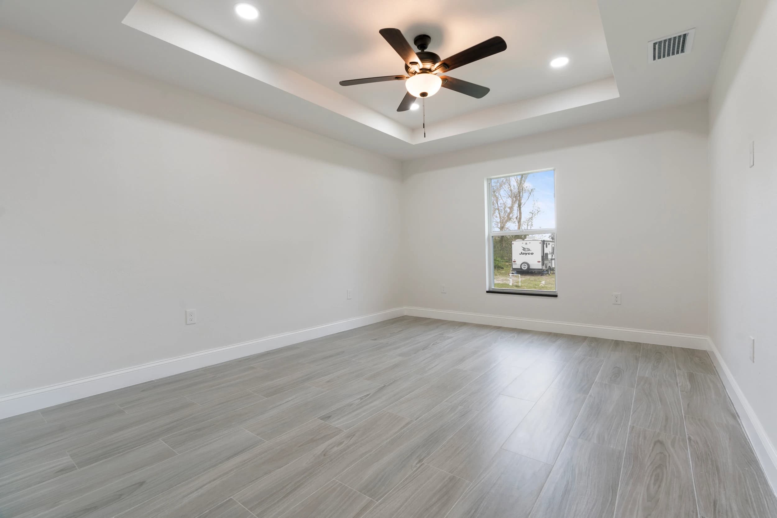 Spacious primary bedroom in a newly built Florida home by Vreeland Roofing, featuring a tray ceiling with recessed lighting, a ceiling fan, and a large window providing natural light.