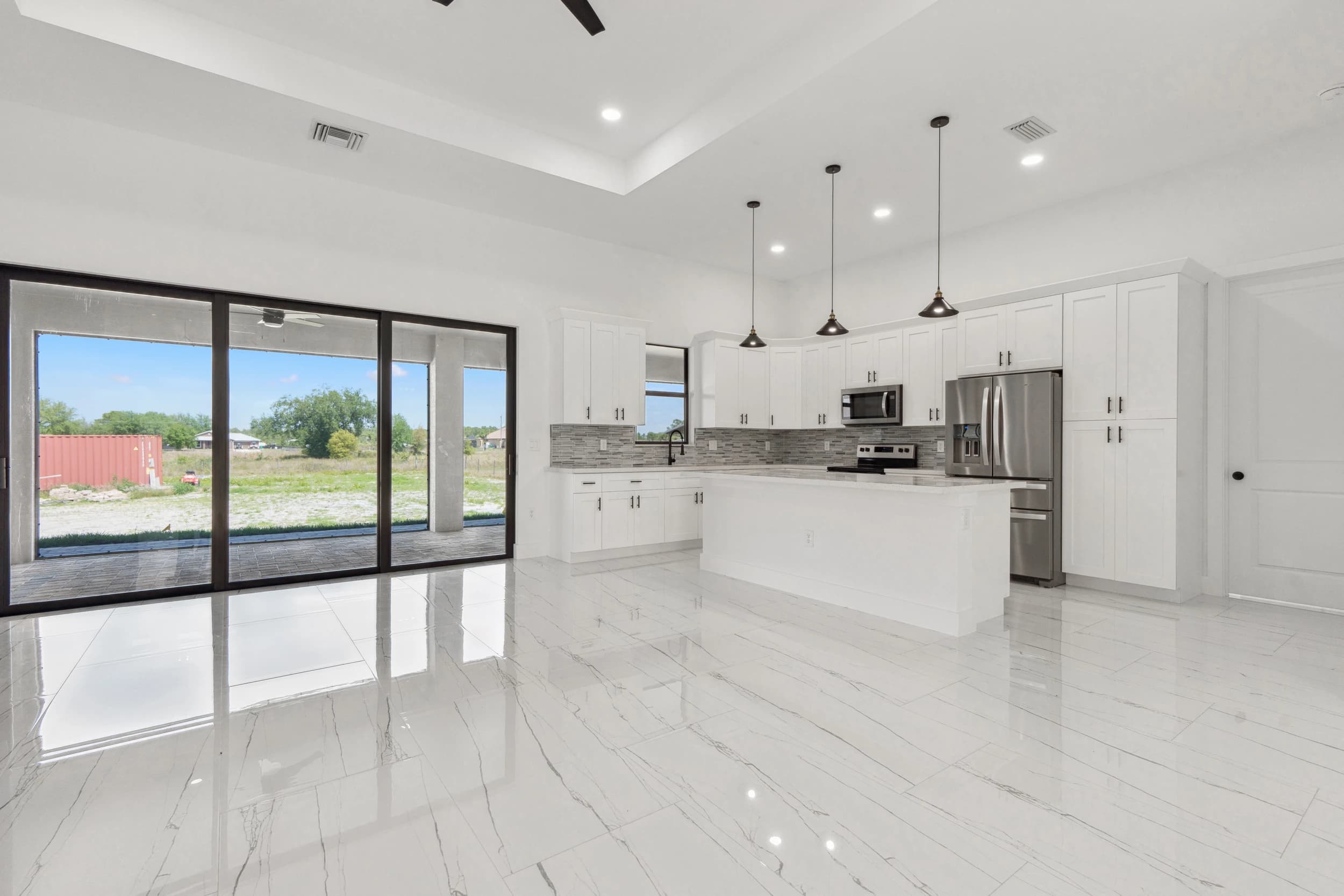 Modern kitchen renovation featuring white cabinetry, stainless steel appliances, marble-style flooring, and pendant lighting. Open-concept design with large sliding glass doors providing natural light and outdoor access. A Vreeland Roofing home remodeling project in Florida.